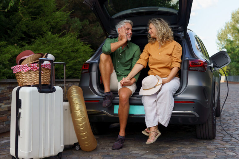 A middle-aged couple sitting in trunk while waiting for charging car before travelling on summer holiday.